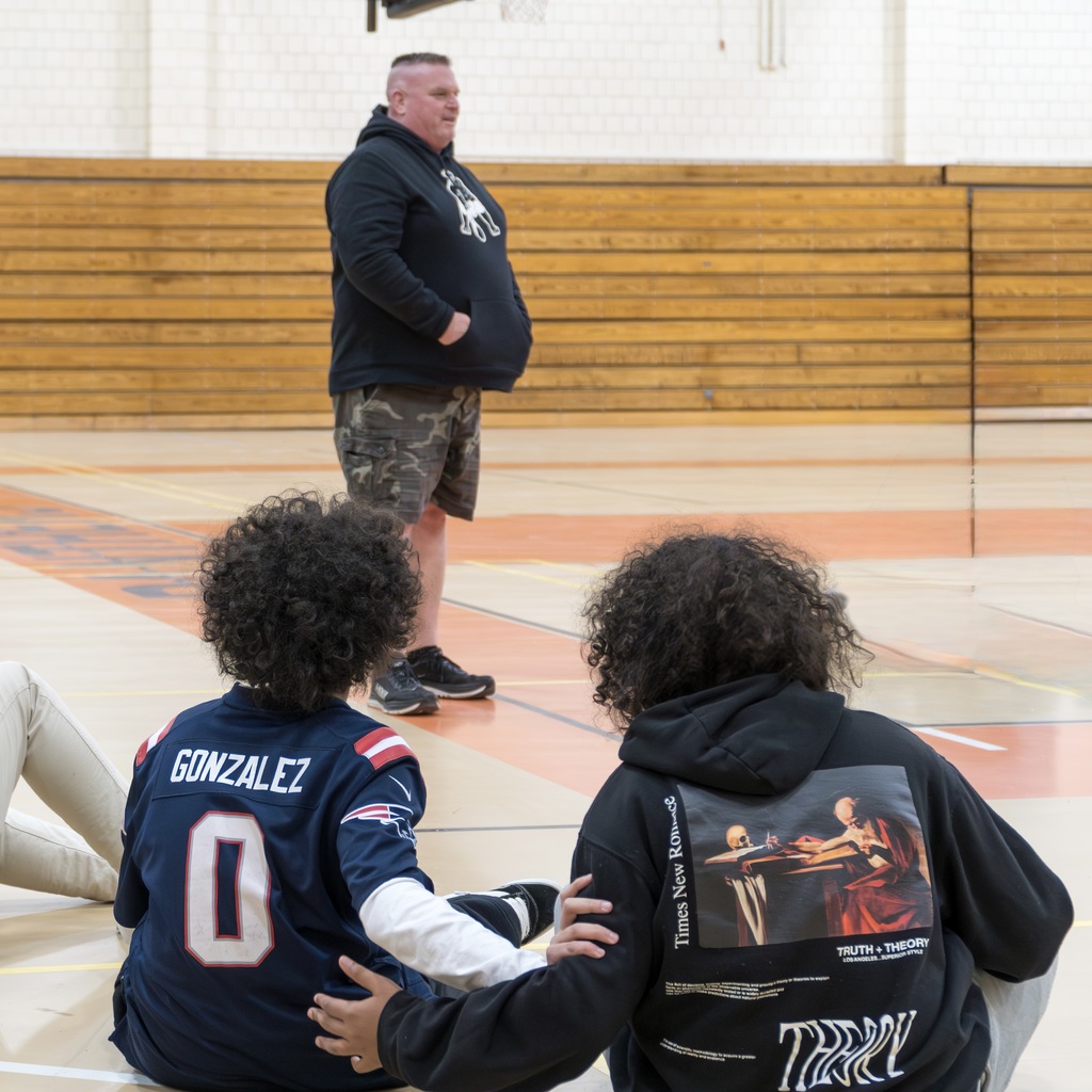 A teacher wearing a Patriots hoodie stands in the middle of a circle of MHS students sitting down in the gym. One of the students wears a Patriots jersey.
