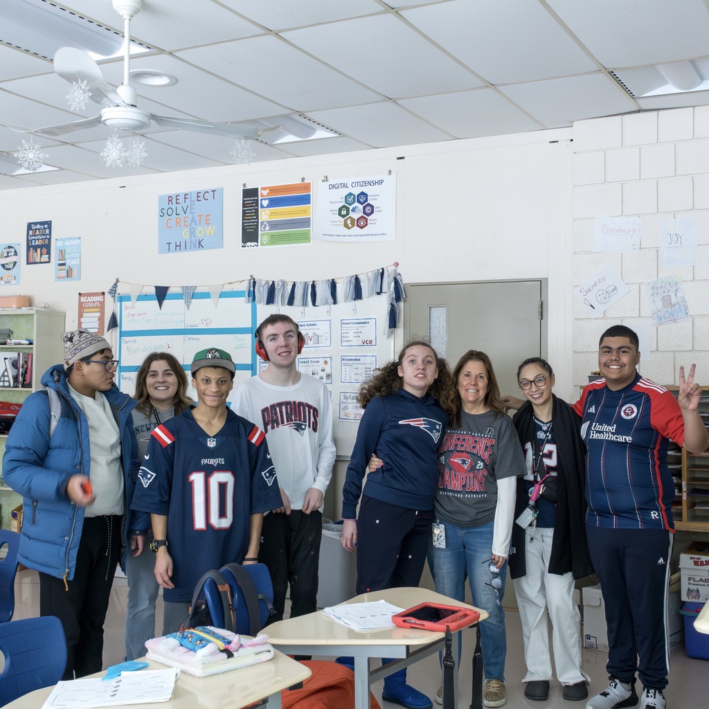 A group of students and their teachers dressed in Patriots gear stand for a group photo in their classroom.