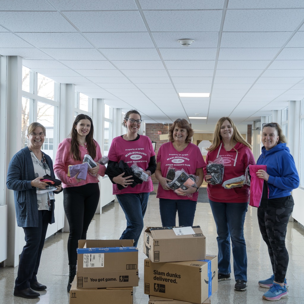 A group of women in pink shirts stands in a hallway with boxes, all are holding socks or scarves.