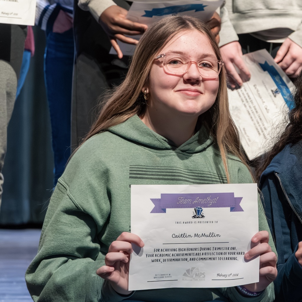 A young female student in a green hoodie sits on stage holding a certificate. Behind them, a group of students stands holding certificates.