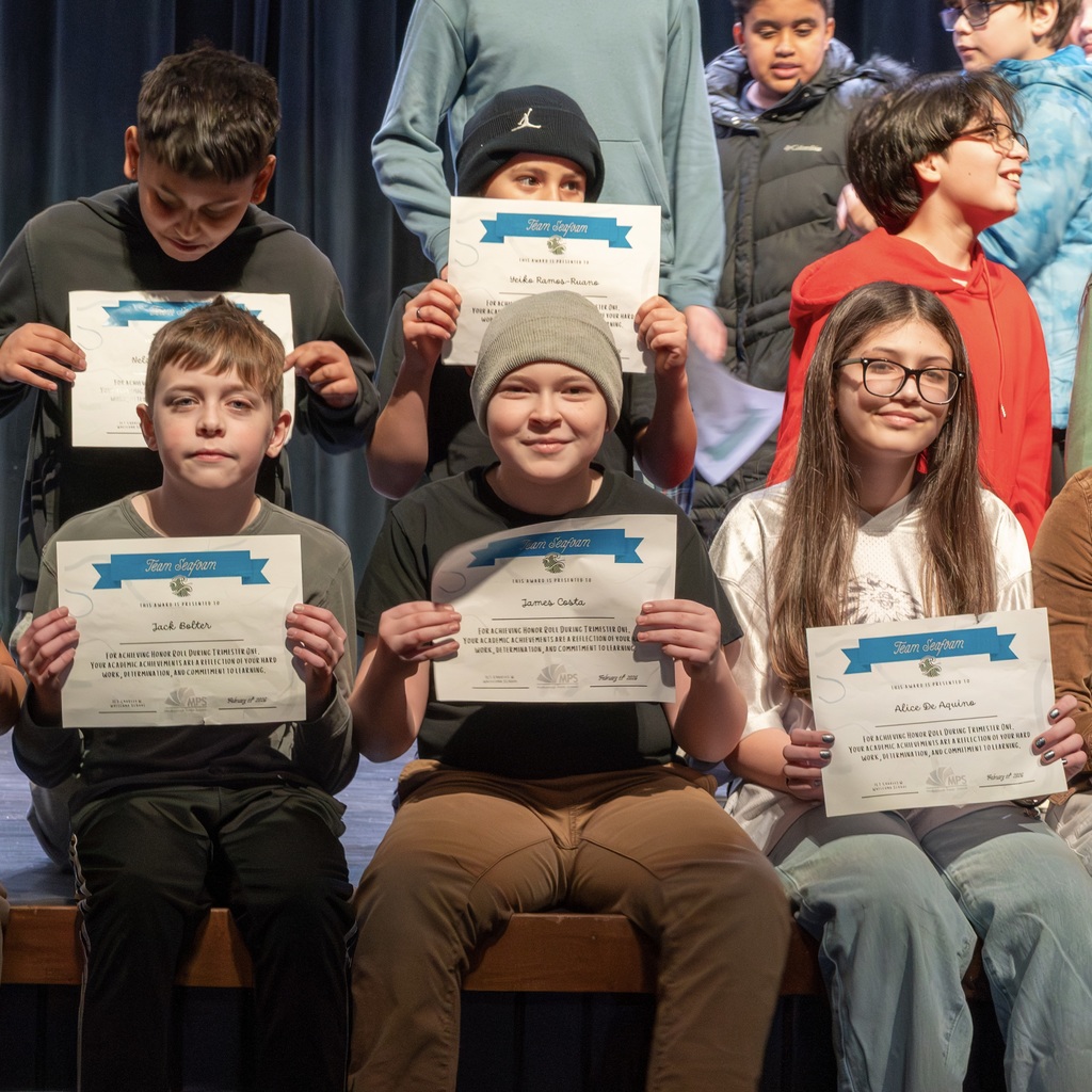 Students hold certificates. Some wear glasses. They sit on a stage. The background shows more students standing or kneeling.