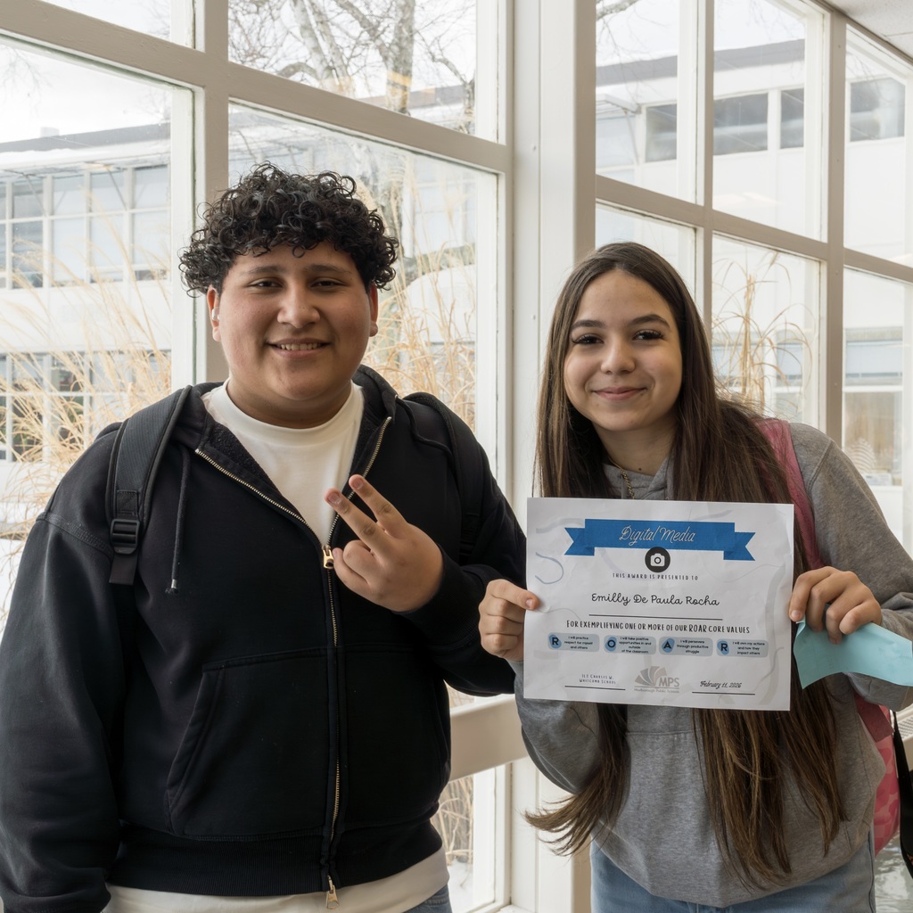 Two students stand by a window, one smiling at the camera and the other holding a certificate.