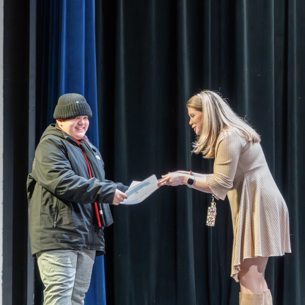 A male student wearing a beanie and a jacket receives a certificate from a female staff member wearing a dress. They stand before a curtain backdrop.