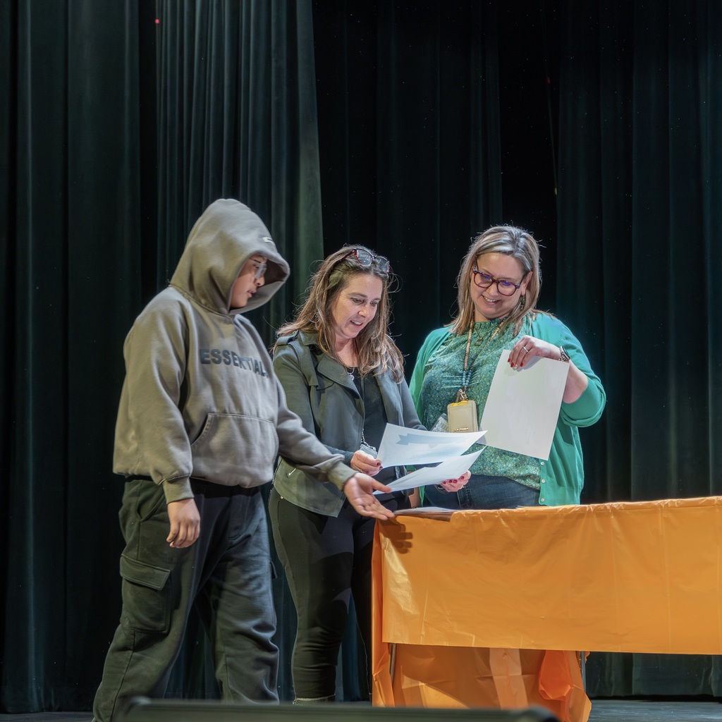 Two female staff members and a student are on stage near a table, holding and looking down at some certificates.
