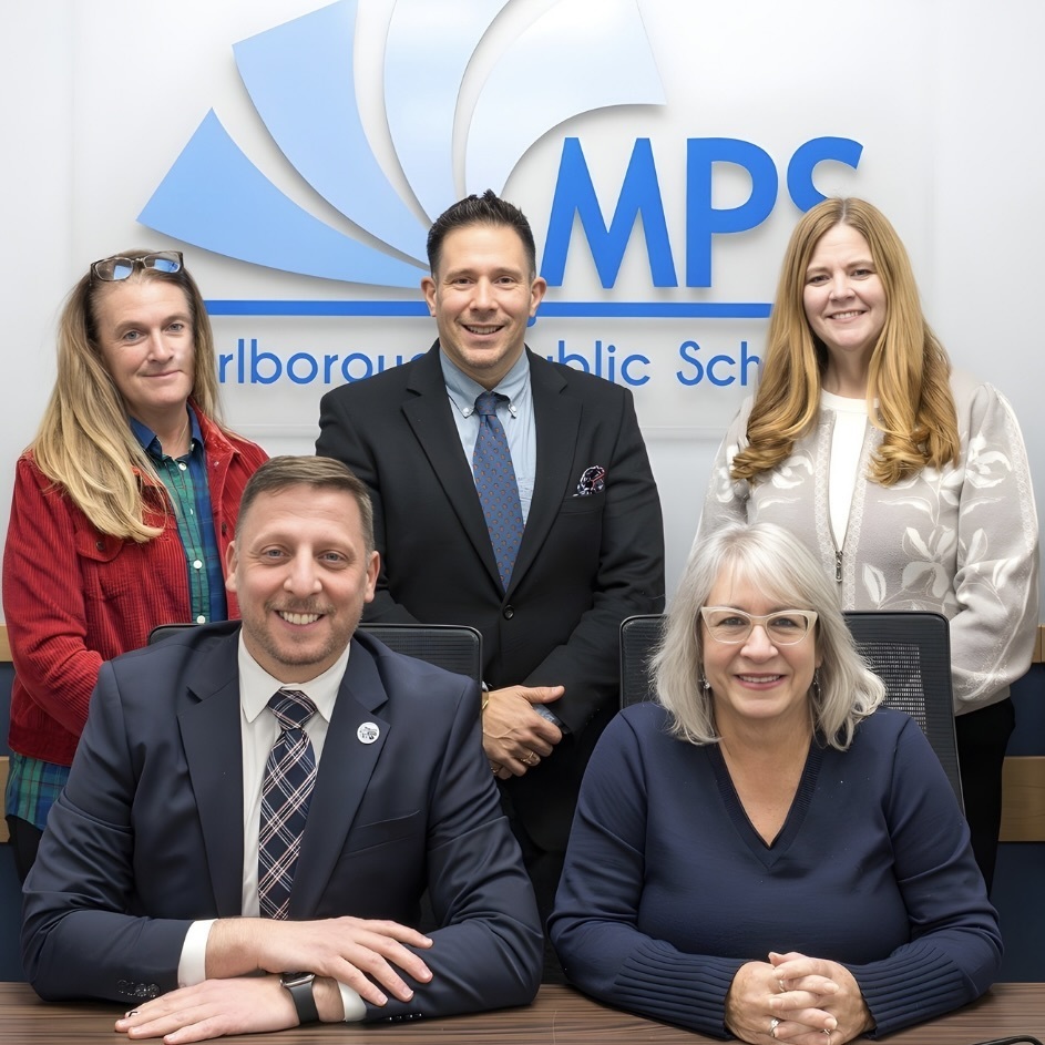 Five Marlbrough School Committee members in business attire sit in front of a sign with the MPS logo.