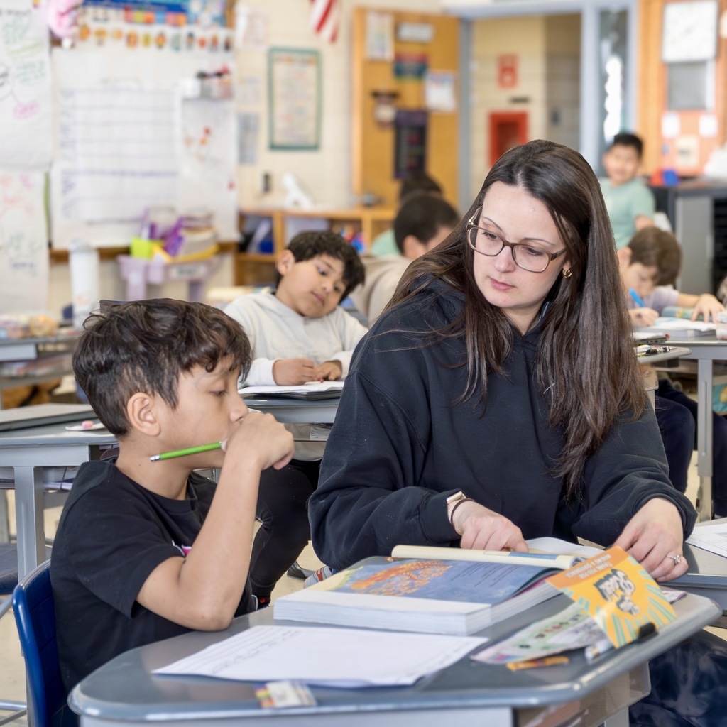 A daily sub sits at a desk next to a student to help him complete a writing assignment.