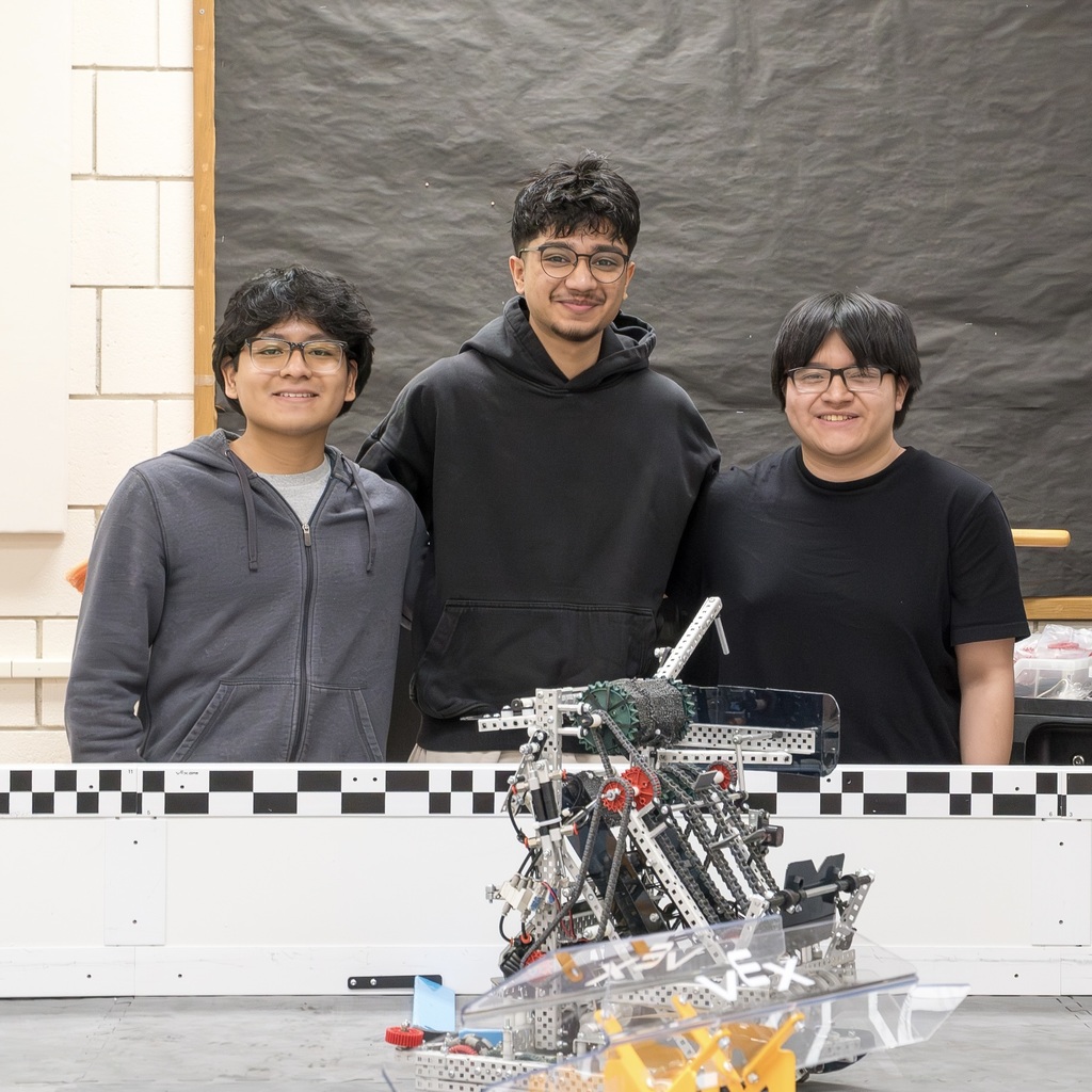 Three MHS Robotics Team members, two in glasses, stand behind a robot on a checkered platform, with a black backdrop.