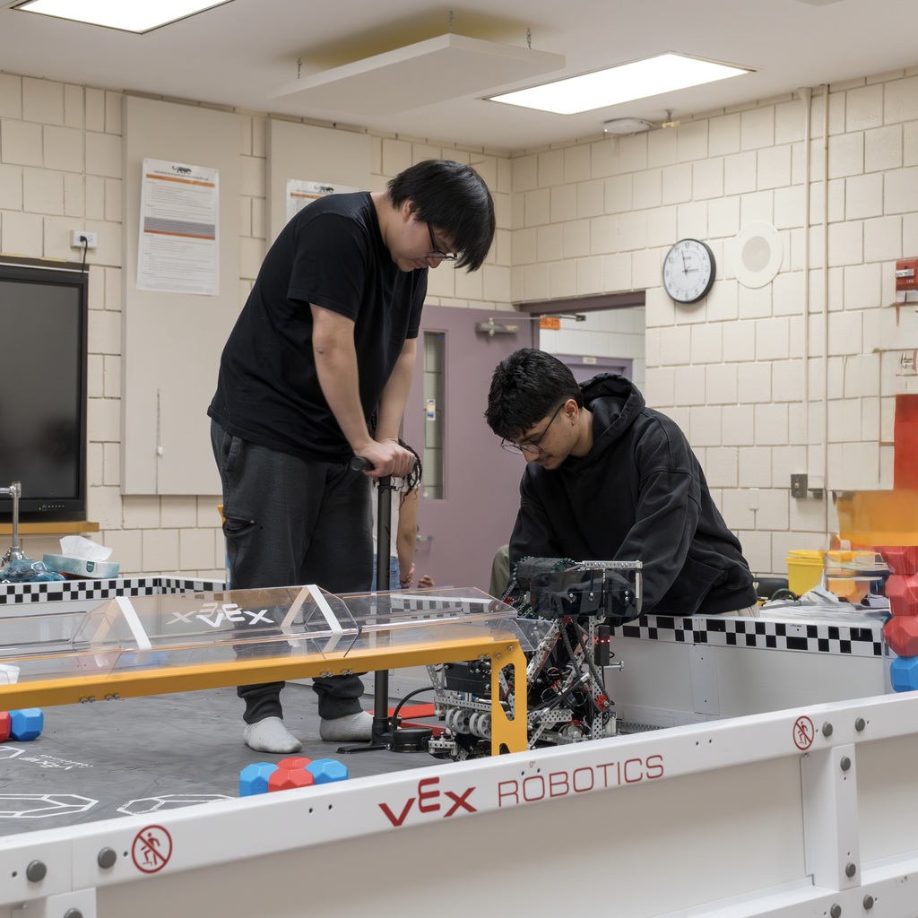 Two MHS Robotics Team members in a classroom, one holding an air pump and the other operating a robotic device.