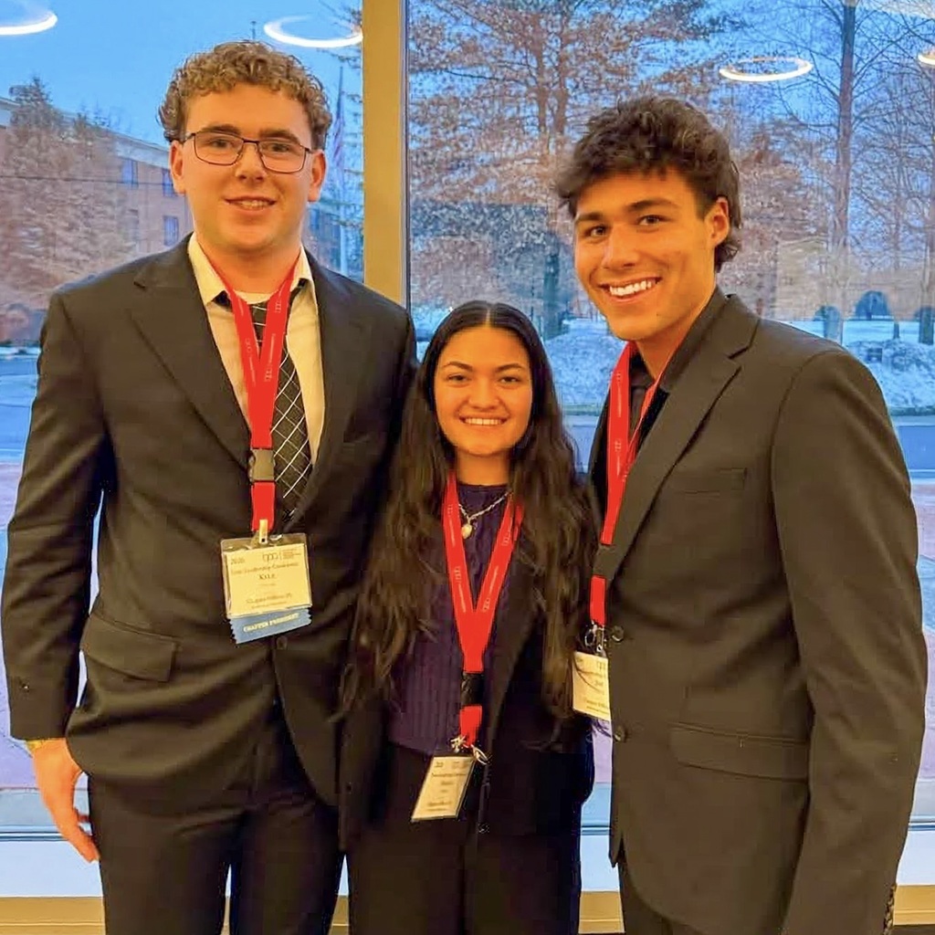 Three MHS BPA CLUB members, two boys and one girl, in suits and lanyards with badges stand together in front of a window.