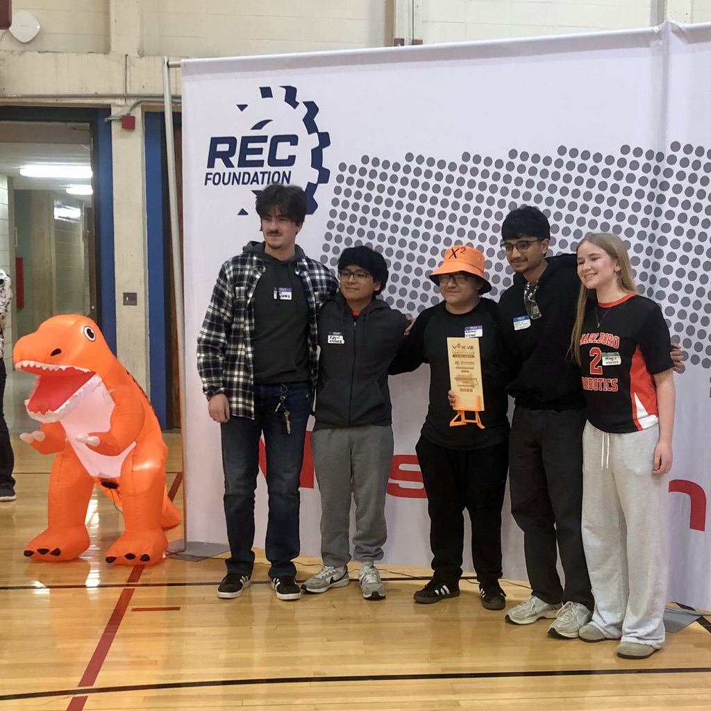 Five MHS  robotics team members stand in a gym with a REC Foundation banner, one holding a trophy. A dinosaur mascot is nearby.