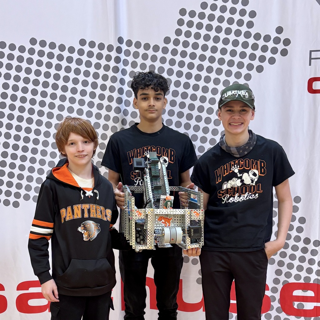 Three young people stand together, two wearing black hoodies and one wearing a baseball cap. One holds a mechanical object.