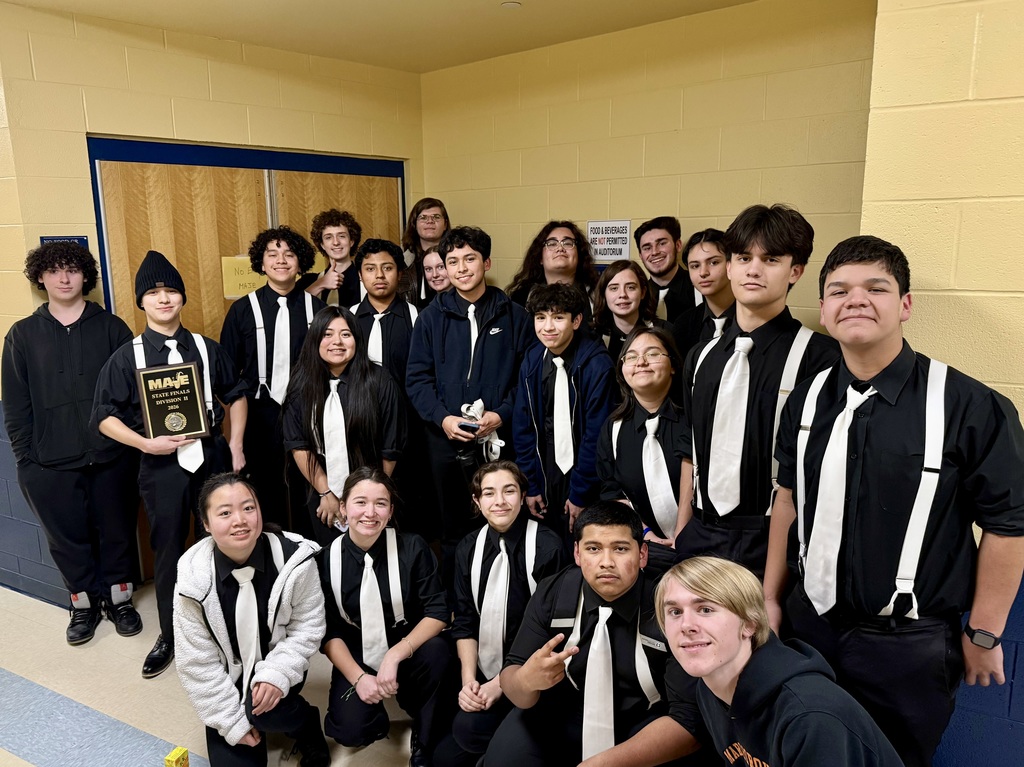 The MHS Jazz Ensemble, wearing black-and-white uniforms and some holding awards, stands together in a room.