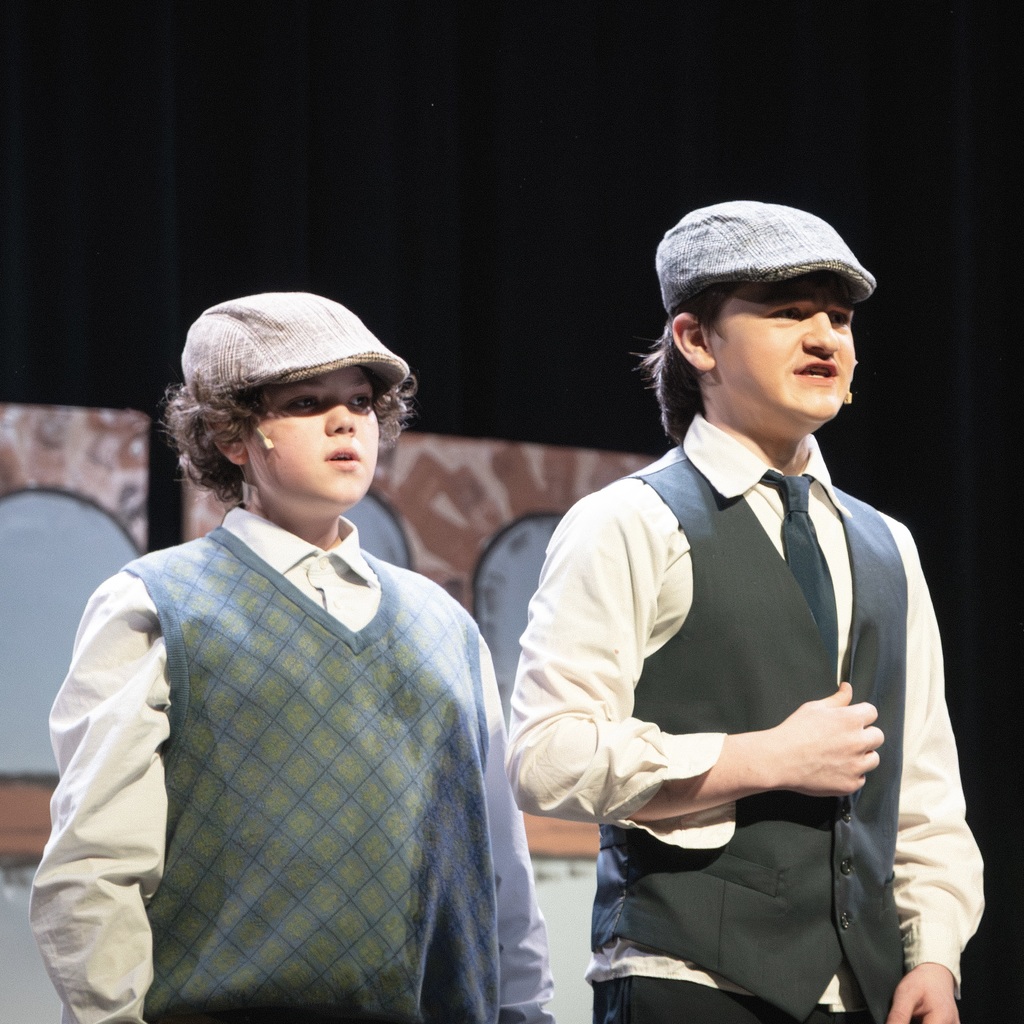 Two young actors on stage, one in a plaid vest and the other in a dark vest, both wearing hats.