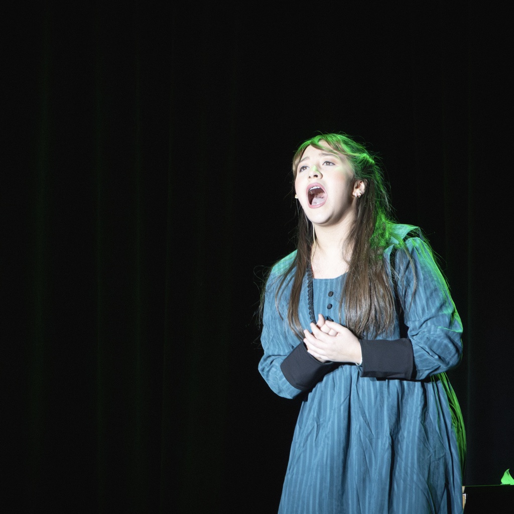 A female student with long hair singing on a stage, dressed in a blue striped dress with navy cuffs.