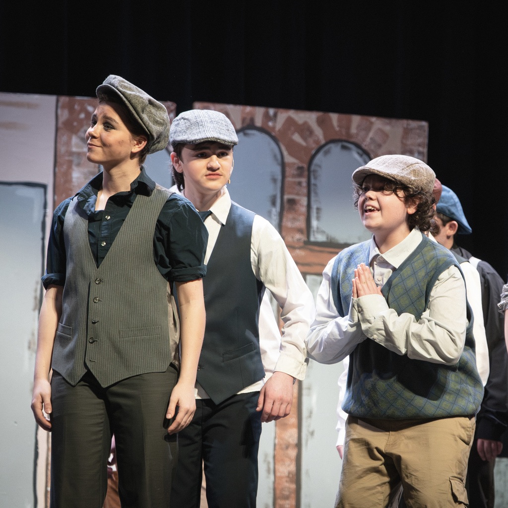 Four Newsies Jr. cast members in hats and vests stand on a stage with brick walls in the background.