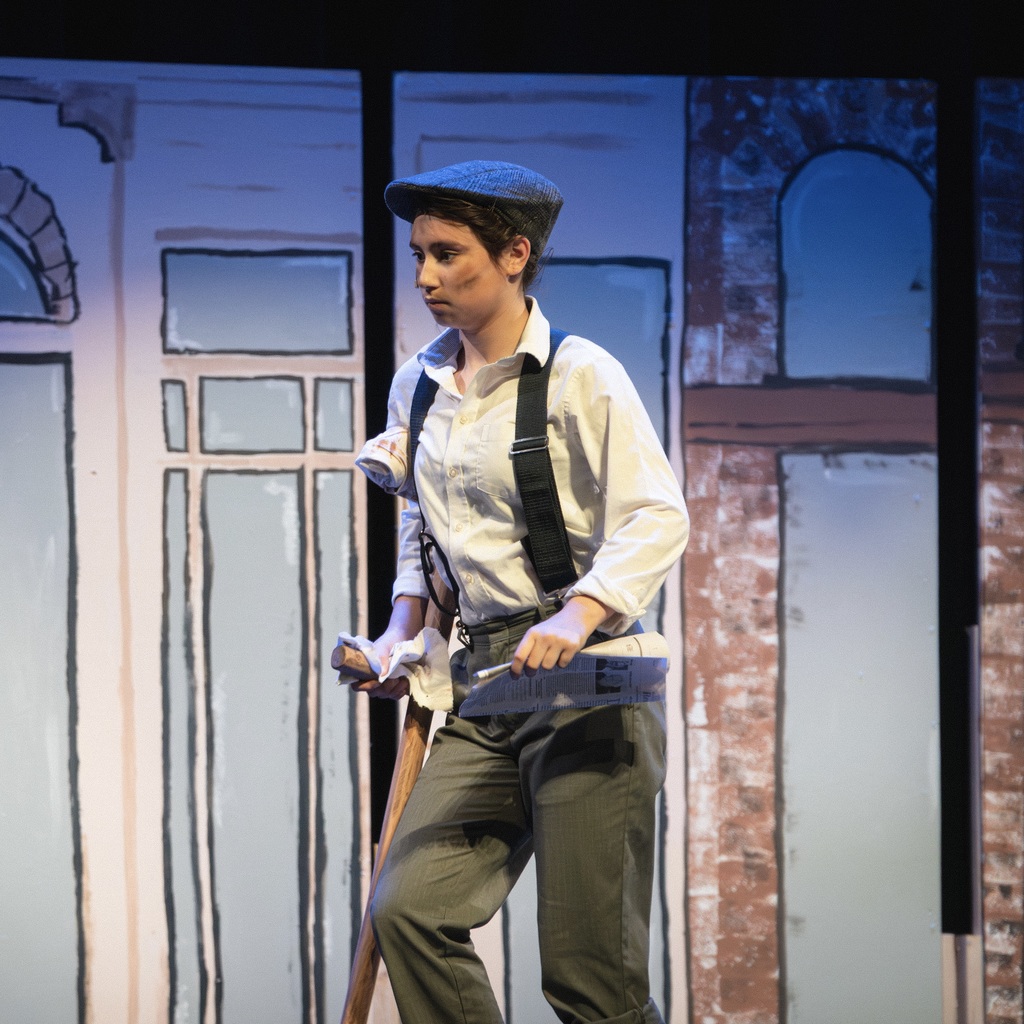 A student actor in a hat and suspenders holds a newspaper and walks with a crutch on a stage with a drawn brick wall background.