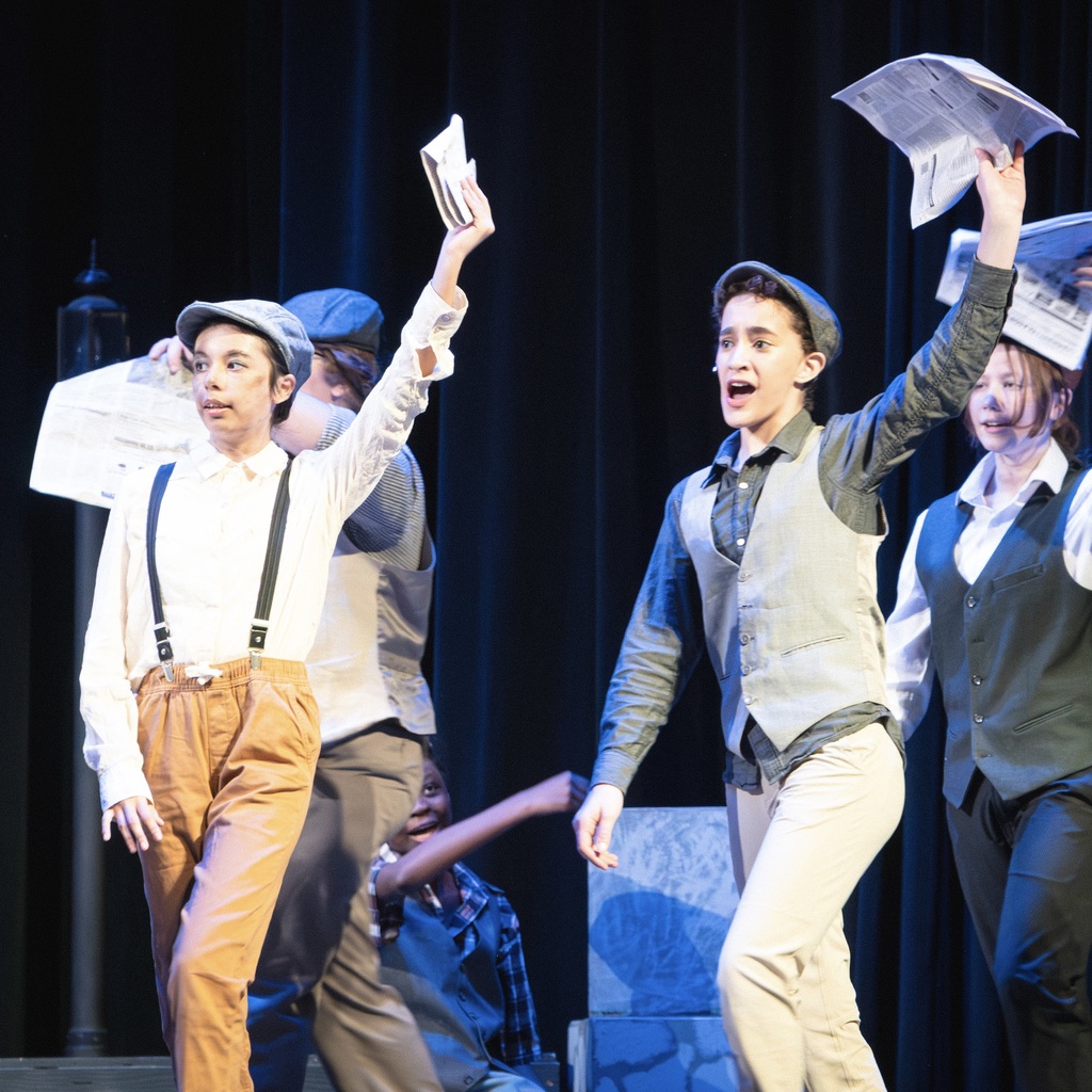 Five cast members on a stage wearing vintage clothing. Four of them stand and hold up newspapers.