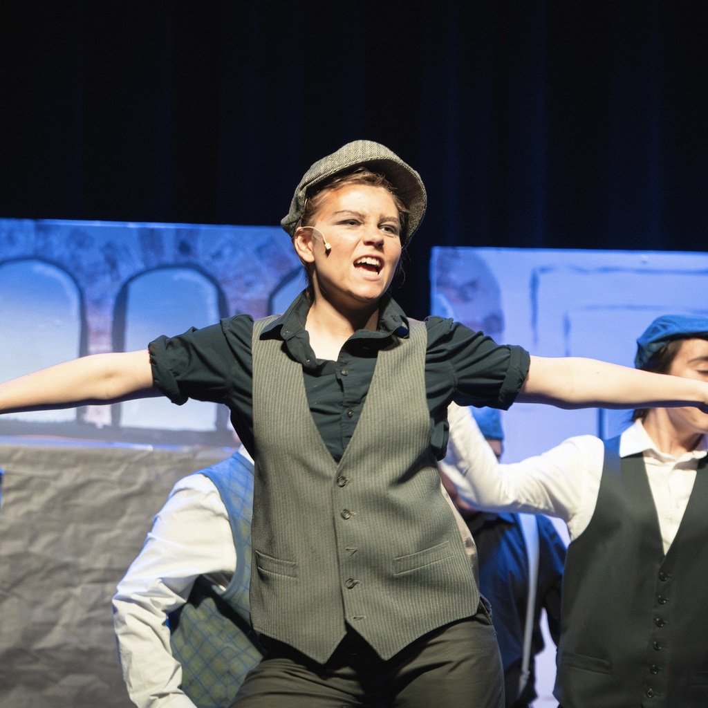 A cast member performs wearing a hat, a vest, and a black shirt, arms raised on stage, with two other student actors behind.
