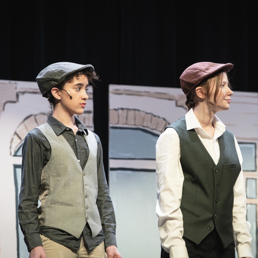 Two Whitcomb Musical Troupe students, dressed in hats, vests, and button-down shirts, on a stage. A mural is behind them.
