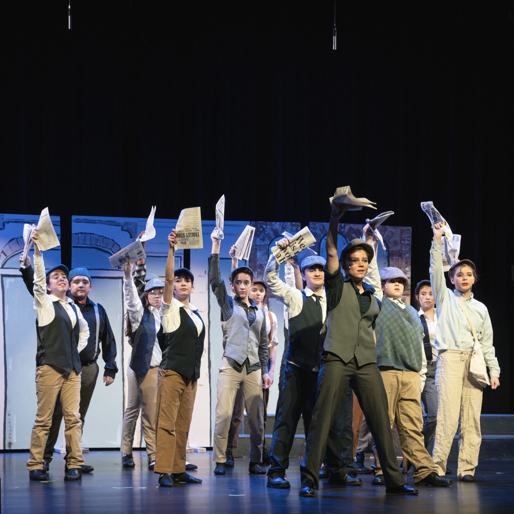 A group of students on stage holding up newspapers, performing Newsies, Jr.