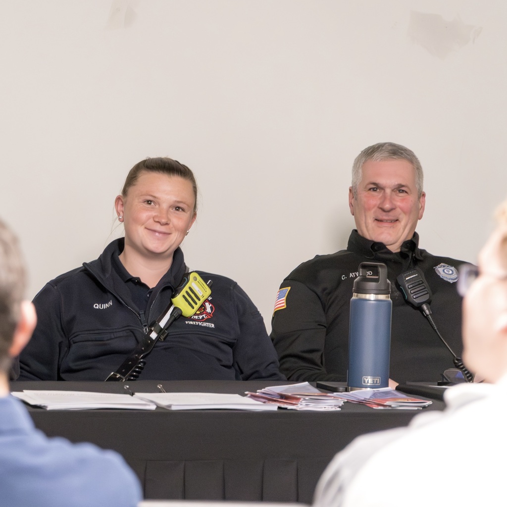 Two first responders, in uniform, sit at a table, smiling directly at the camera.