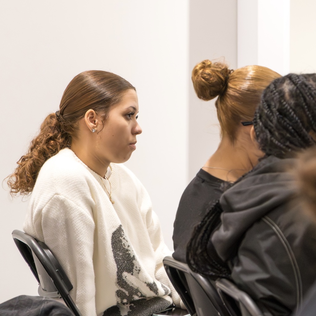 Three female MHS students are seated in chairs. One student in white listens intently, looking ahead, with two others on her right.