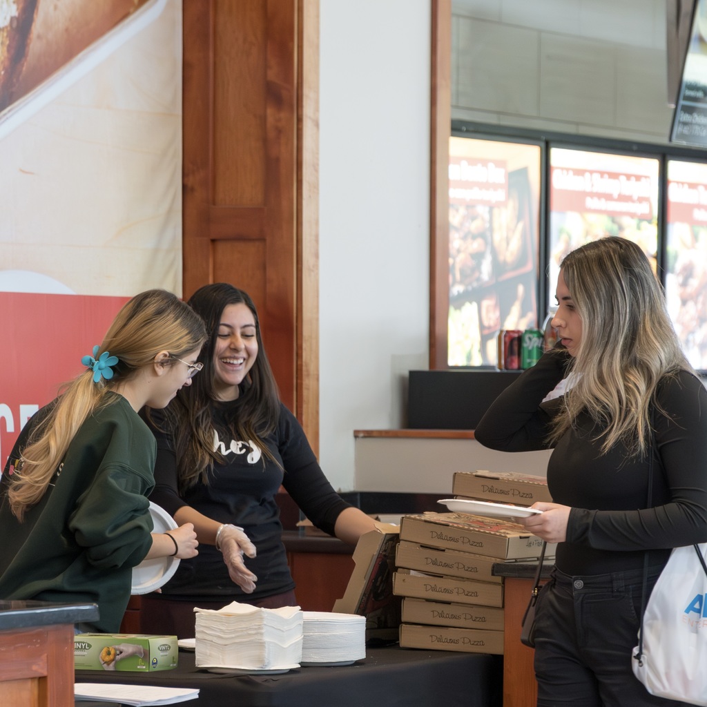 Three women in the Solomon Pond Mall food court, one in green, another in black, serve pizza to the third, who is holding a plate and a white bag.
