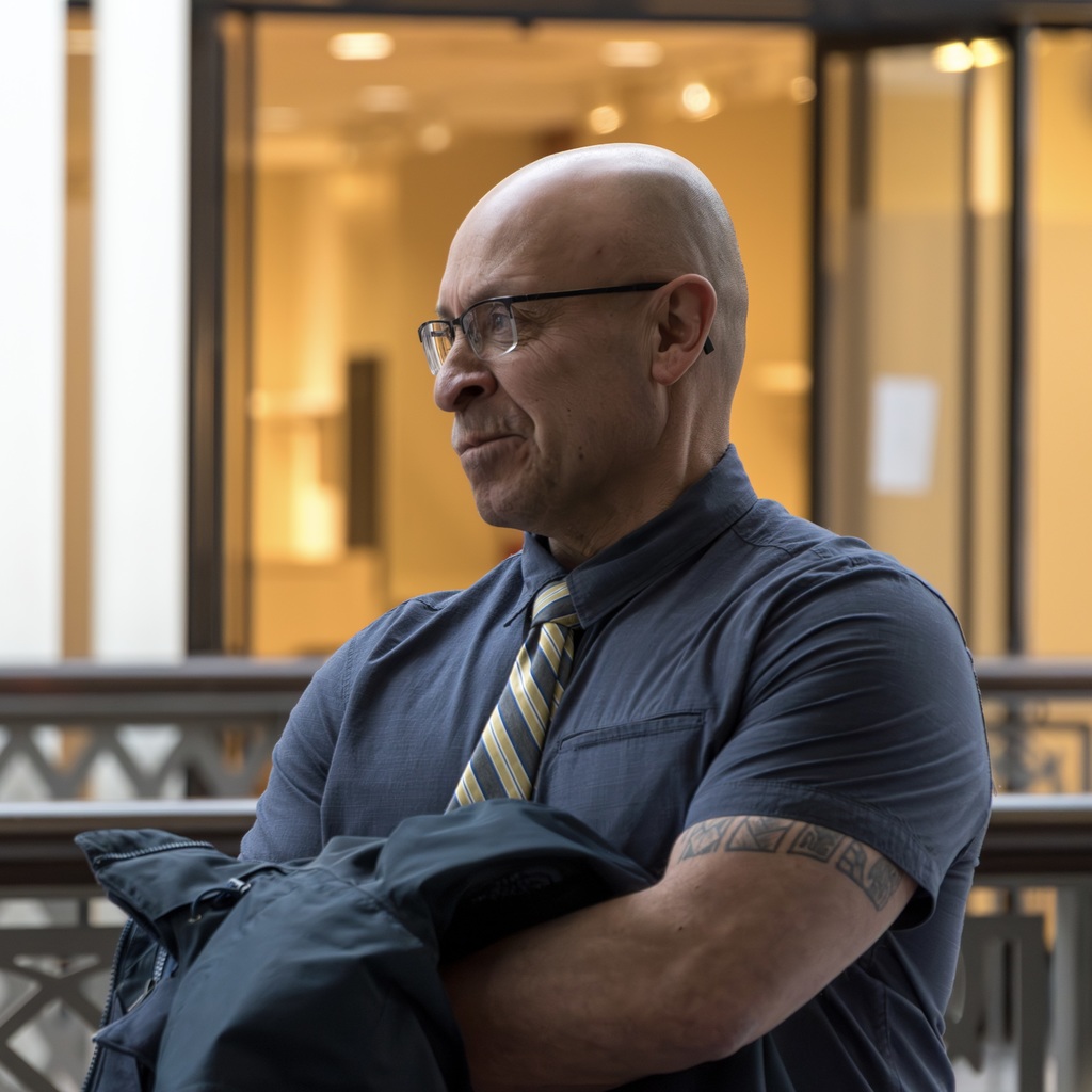A male MHS staff member with glasses and a striped tie holds a coat over his arms, looking to the side.