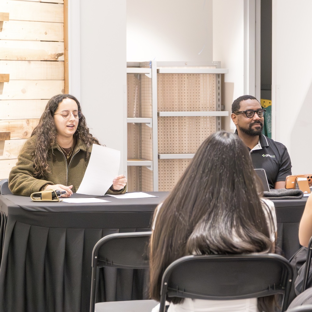 Two individuals sit at a table, one reads while the other listens. In front of them, a student sits on a chair.