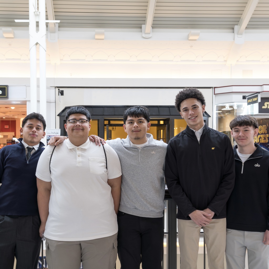Five male MHS students stand together inside a mall. They wear various colored shirts and pants. Behind them are shops with signs.
