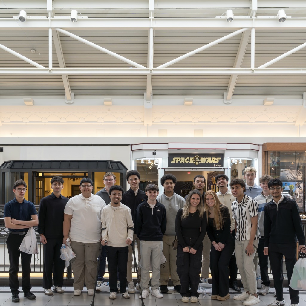 A group of MHS students poses on the second floor of the Solomon Pond Mall. They wear casual clothes, some holding bags.