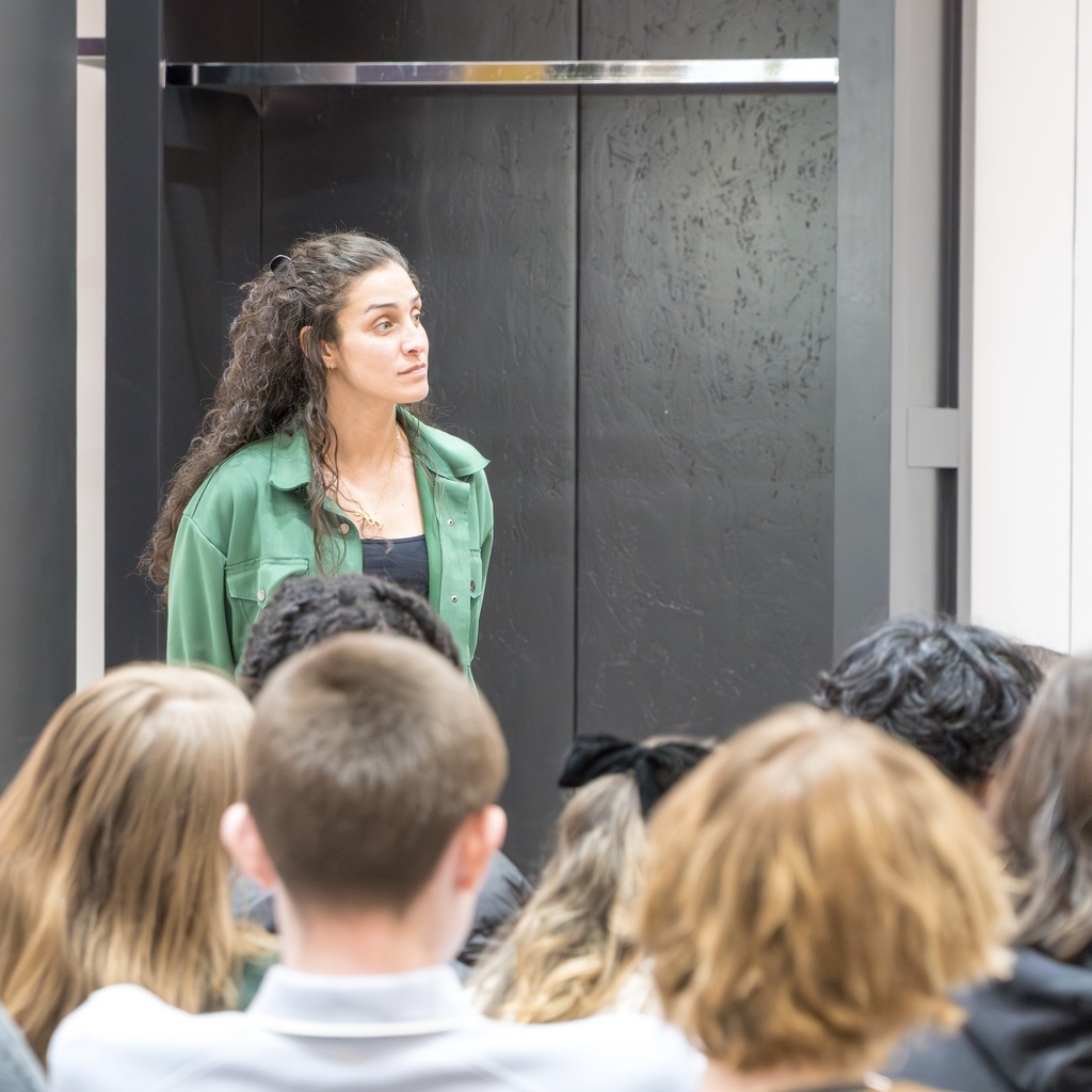 A  female MHS staff member with curly hair stands in front of a group of students in a room with a black background.
