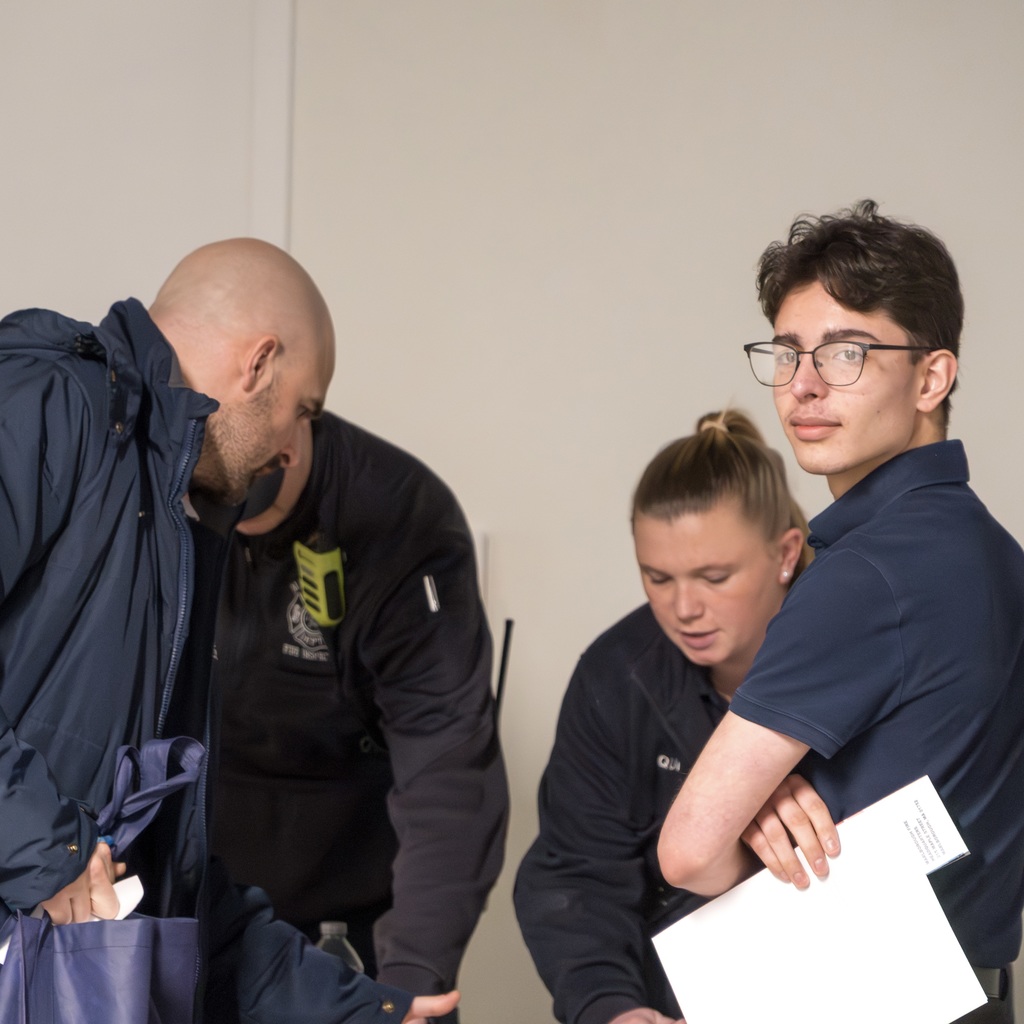 Two first responders in uniforms stand next to a light-colored wall. A man on the left faces them, holding a blue bag. An MHS student stands on the right, holding a white sheet of paper and looking directly into the camera.