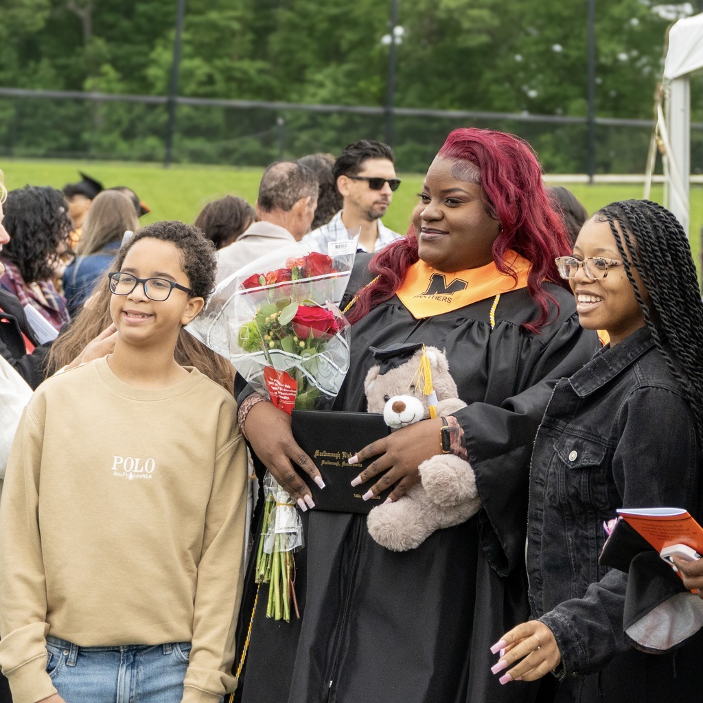 An MHS female graduate with red hair holds flowers and a stuffed animal, standing with a young boy and another woman, all outside.