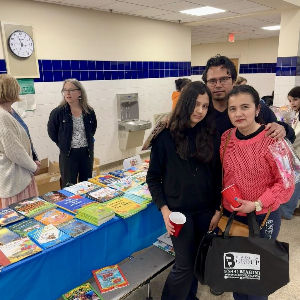 A group of people poses at a table with books, a blue tablecloth, and a clock on the wall.