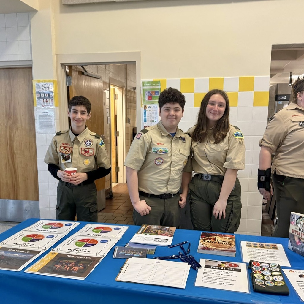 Three youths in scout uniforms stand at a table, with papers and books spread out. A yellow-and-white checkered wall is in the background.