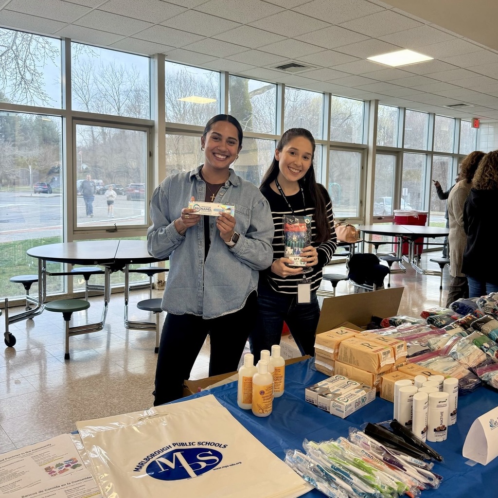 Two women standing in a room with tables, smiling and holding items. Behind them are windows and a road.