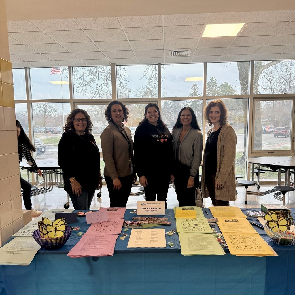 Five women stand behind a blue tablecloth, displaying brochures and papers, in a room with large windows.