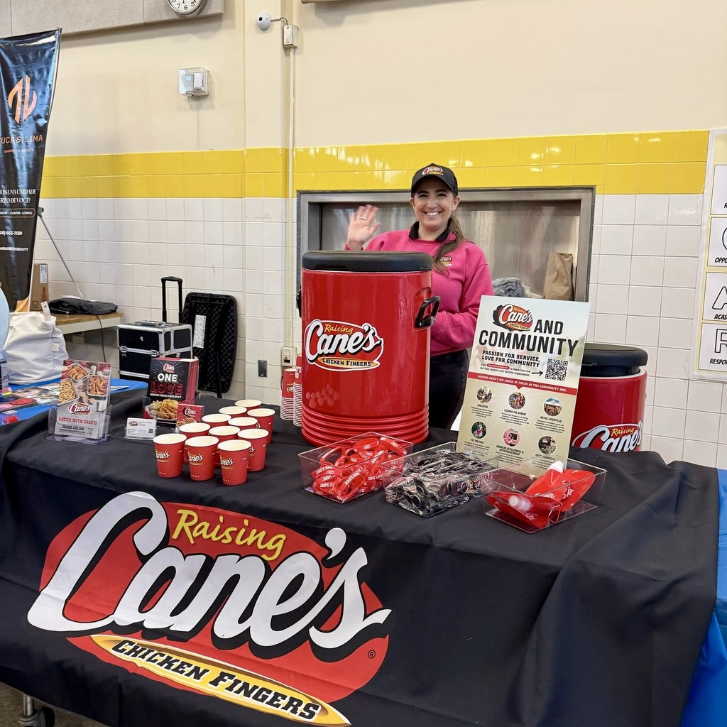 A Cane's employee stands behind a table covered with a black cloth and the Raising Cane's logo. The table has red cups, a large red container, and other items.