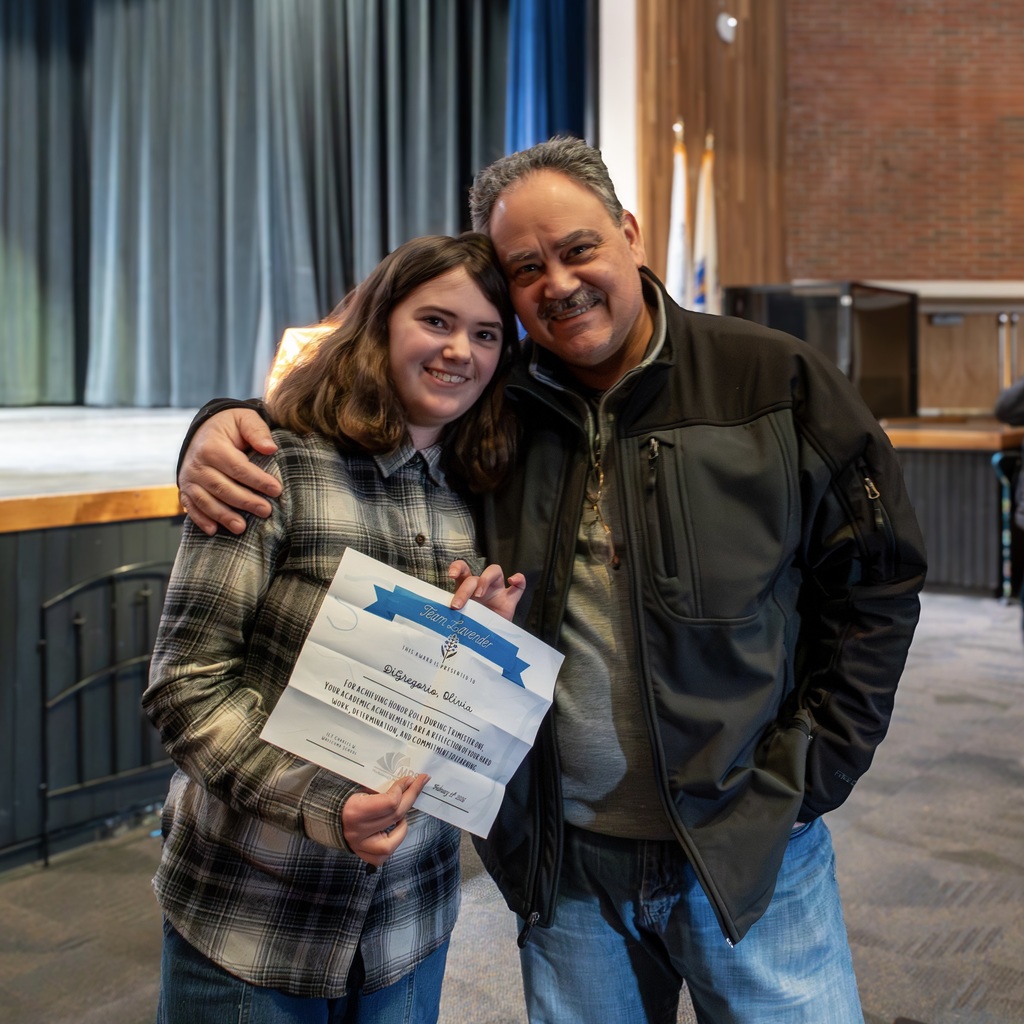 A Whticomb male family member and  student stand together, smiling and posing with a certificate in a room.