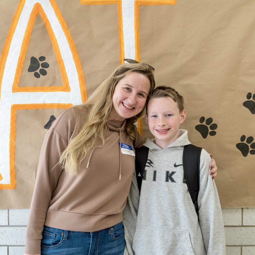 A Whitcomb mom and a young boy stand close, smiling, in front of a wall. The wall features a banner with paw prints and the letter "A".