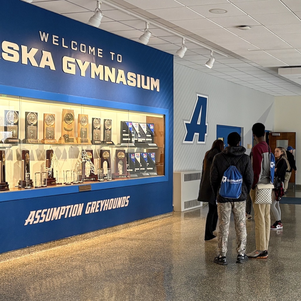 Blue gymnasium wall with "Welcome to Laska Gymnasium" and "Assumption Greyhounds" text. People standing in the foreground.