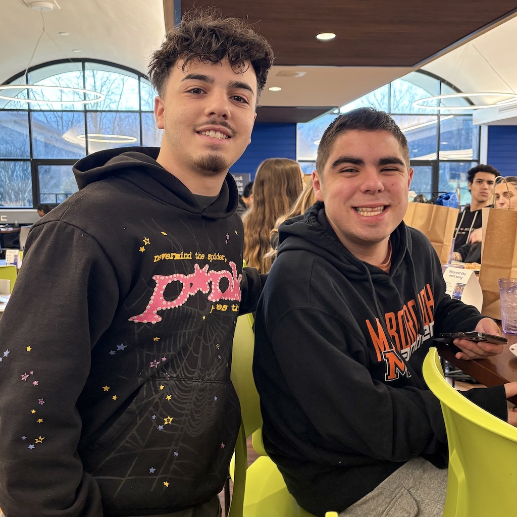 Two young men in black hoodies, one with a spider web design and the other with text. One is standing next to the other one who is sitting at a table.
