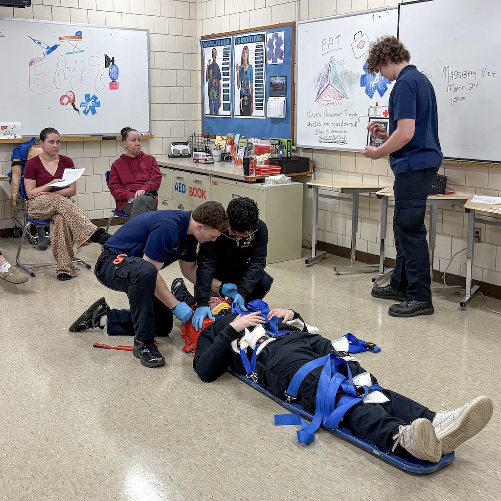 A group of students wearing blue uniforms practice first aid on a mannequin in a classroom.