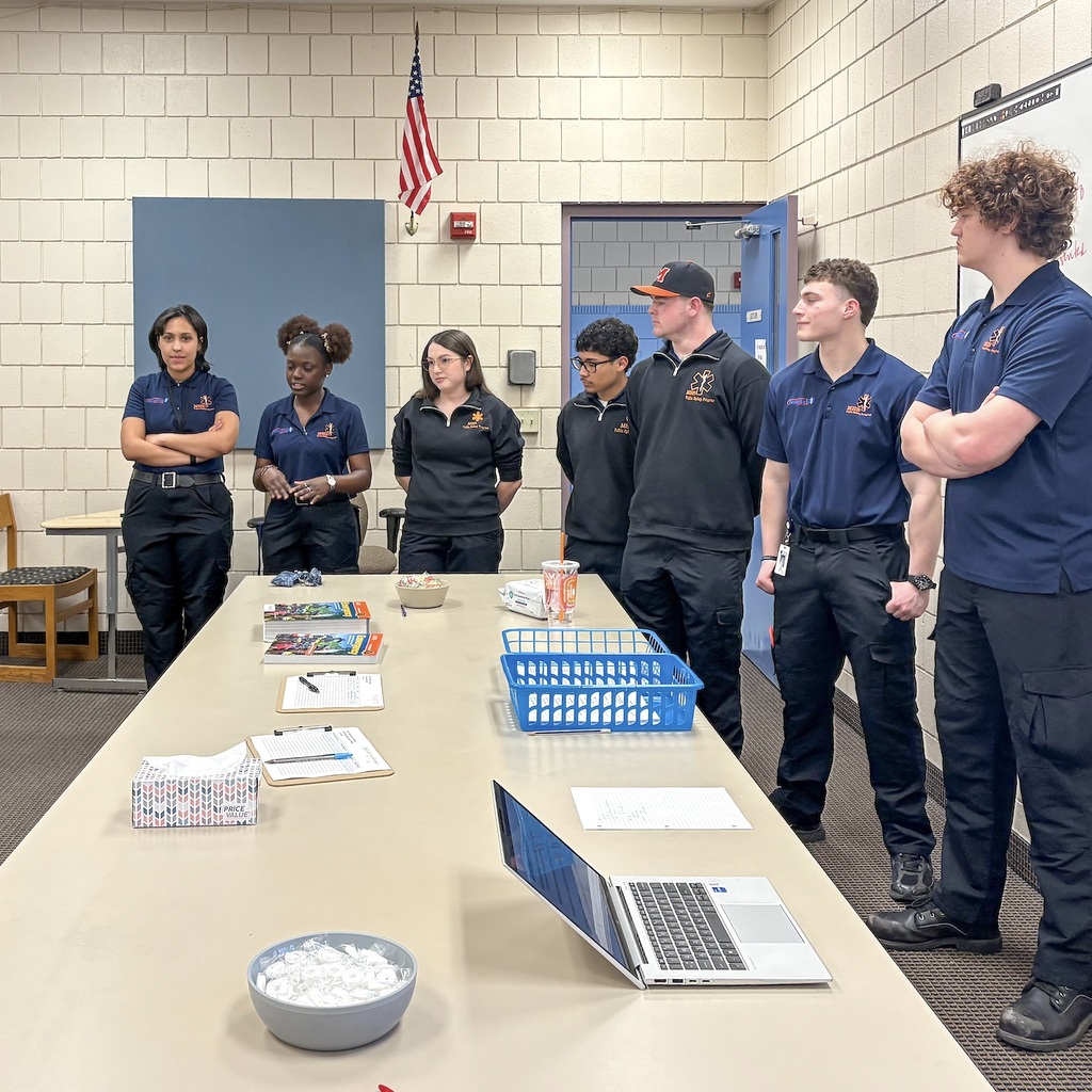 A group of people in uniforms stand around a table with a laptop, papers, and a bowl.