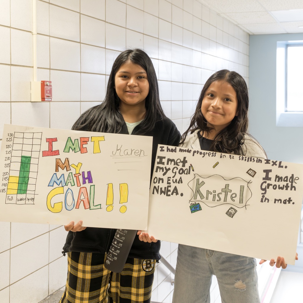 Two girls stand in a corridor holding white signs, one with "I MET MY MATH GOAL!!" and the other with "I made progress in math."