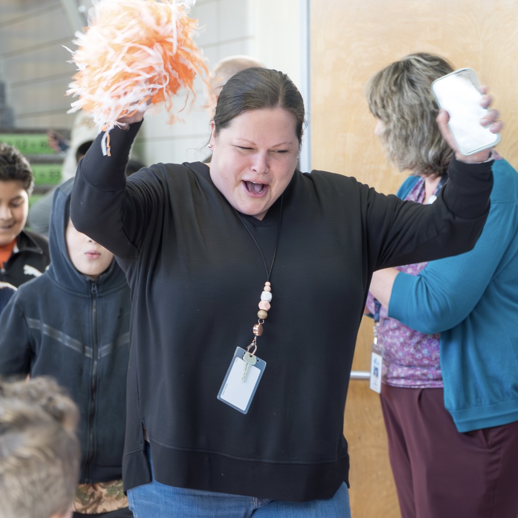 A Richer School staff member with a black top and a beaded ID card, cheering with pom-poms in a school hallway.