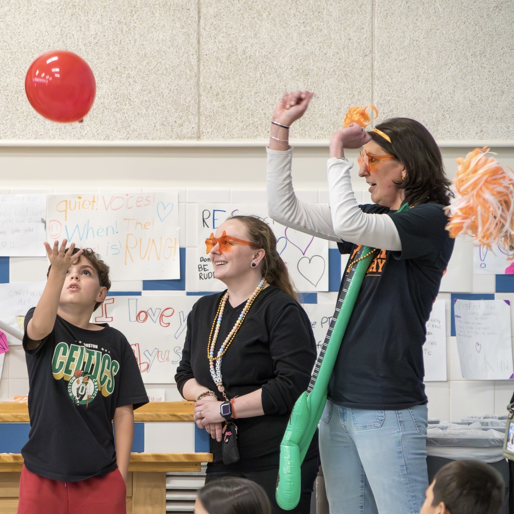Two Richer School staff members and a student are in a room. The staff members wear glasses and cheer on as the student, wearing red pants and a Celtics jersey, plays with a red balloon.