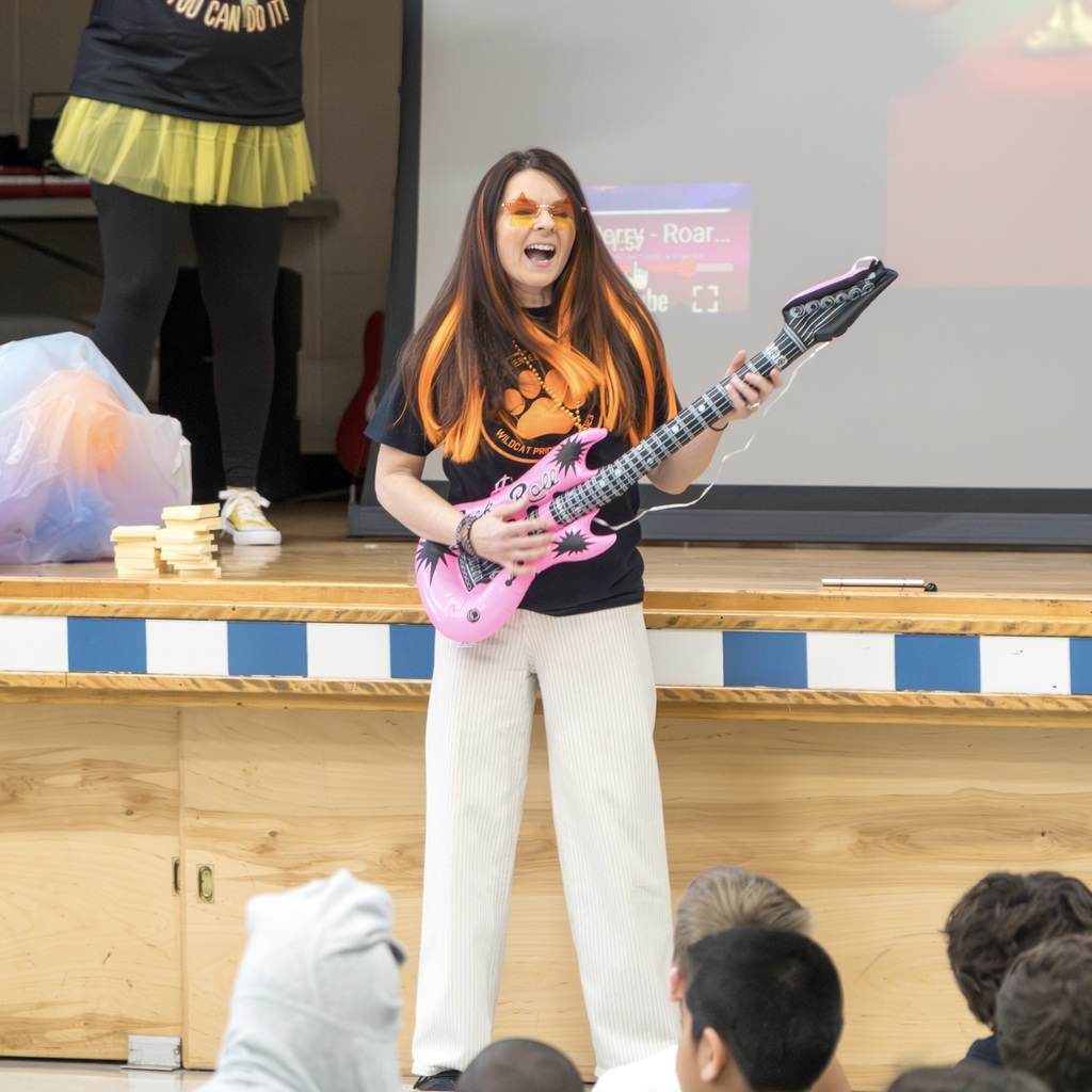 The Richer School principal plays a pink guitar while standing on stage; several students sit in front of her. Behind her, a large screen displays text.