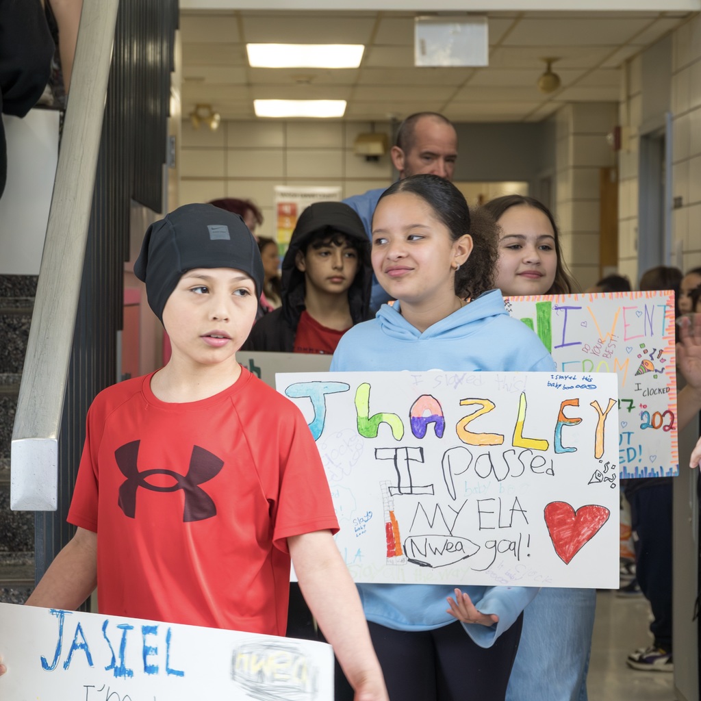 Richer Schools students hold signs with words and illustrations as they walk down a hallway. A boy wears a red Under Armor t-shirt.
